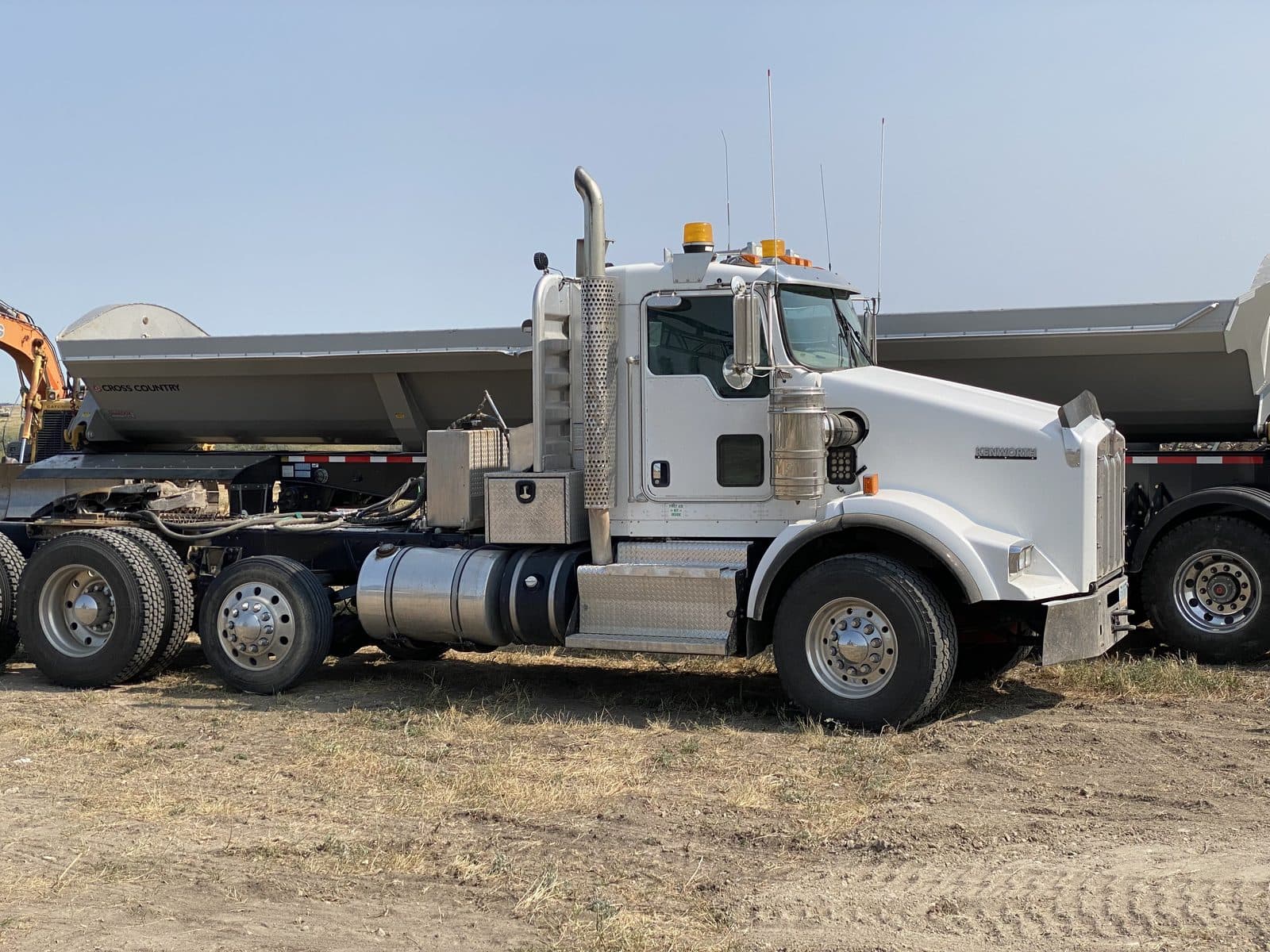 A white Kenworth tractor between two side-dump trailers, parked on the yard