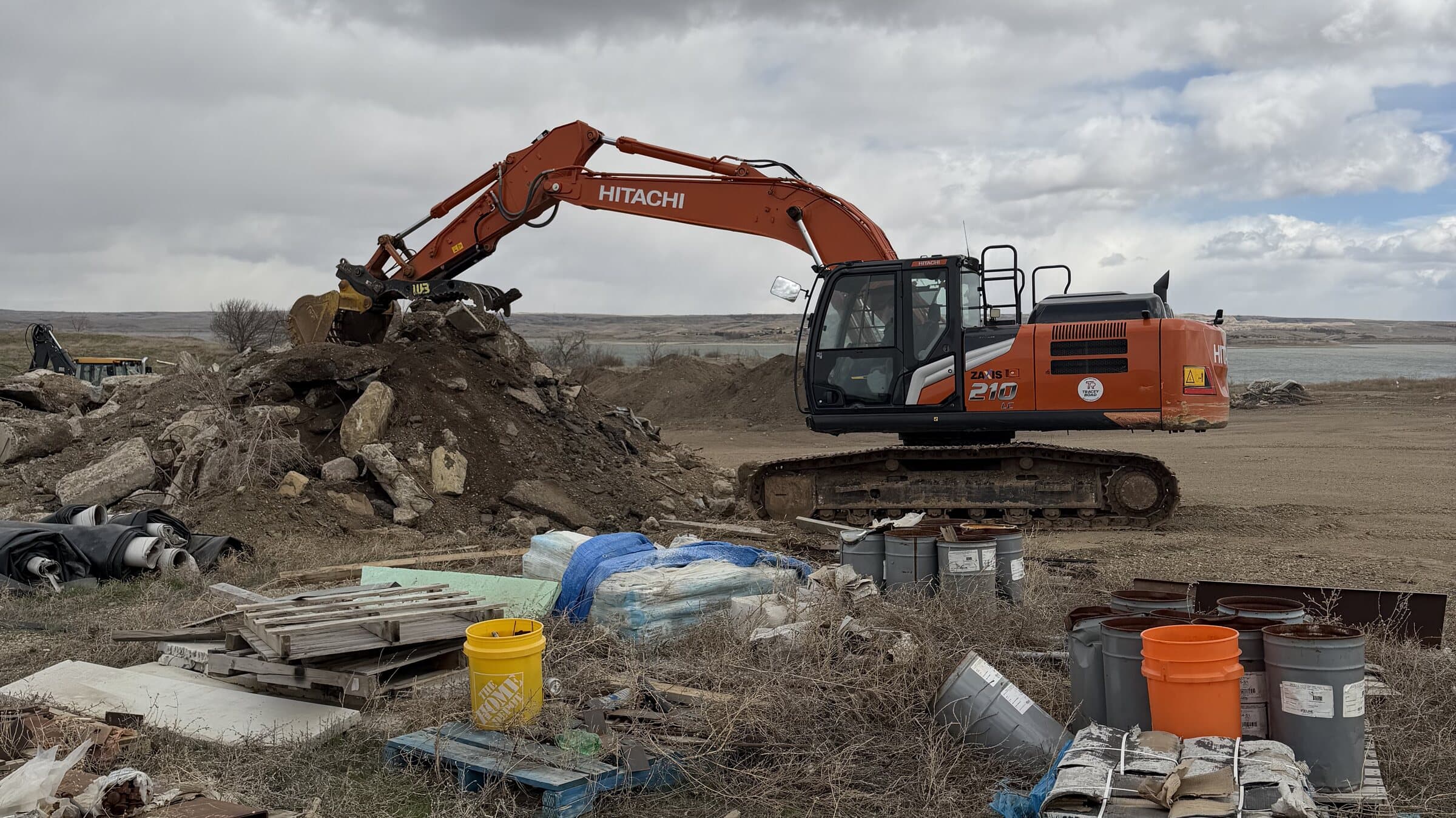 A tracked excavator working a pile of broken concrete at the yard, with open water and bluffs behind it